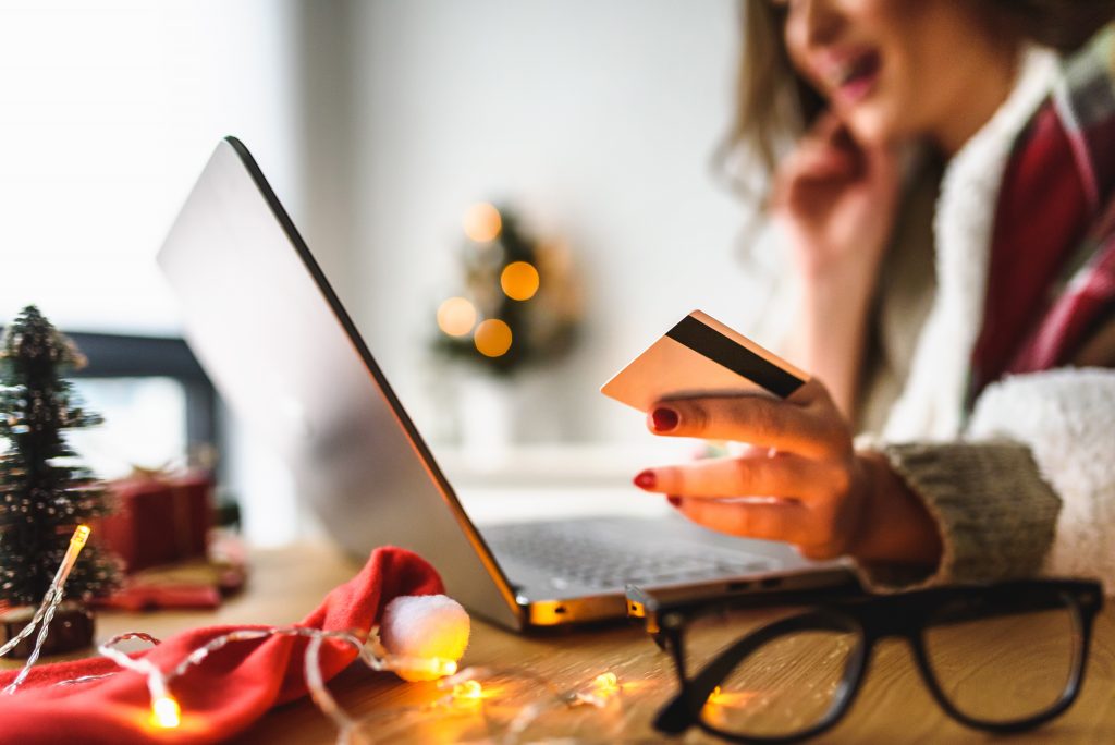 A person is talking on the phone while holding their credit card and using a laptop. Their glasses are in the foreground, and they are dressed for cold weather and there are holiday decorations around them.