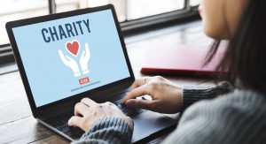 A person by a window using a laptop to access a page that says "Charity" and shows the giving hands and heart. Image licensed through Adobe Stock.