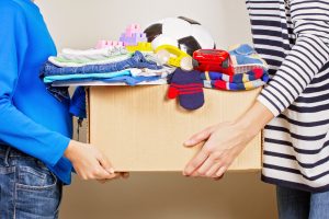 A person in a blue shirt and jeans handing a box of clothing and toys to a person in a striped shirt and jeans, representing direct giving. Image licensed from Adobe Stock.