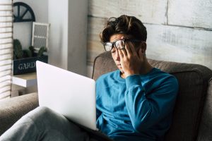 Stressed student at home studying with a laptop and rubbing their eyes. Image licensed through Adobe Stock.