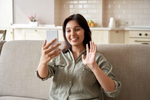 Young indian woman sit on sofa hold mobile phone video calling in app. Image licensed through Adobe Stock.