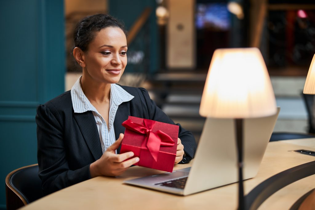 A woman sitting at a laptop with a lamp turned on holding a red ribbon-wrapped package and smiling. Image licensed through Adobe Stock.