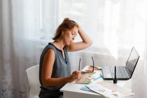 A young woman sitting at her desk, On the table are a laptop and drawings. Image licensed through Adobe Stock.