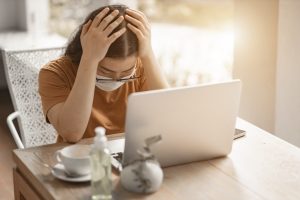 Person seated at desk using a laptop and wearing a protective mask and drinking coffee, holding their head in frustration. Image licensed through Adobe Stock.