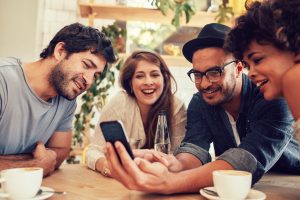 Group of young people sitting in a cafe and looking at the photos on smart phone. Image licensed through Adobe Stock.