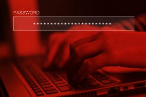 Person typing online account password on laptop computer keyboard with a red filter and the password displayed in an overlay with asterisks above it. Image licensed through Adobe Stock.