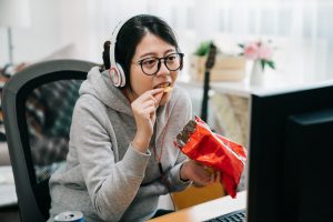 An Asian person sits at home with pack of snacks in his hands eating chips and watching videos on computer screen. Licensed through Adobe Stock.