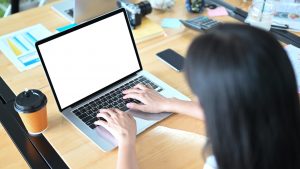 A woman is using a white blank screen computer laptop at the cluttered working desk. Licensed through Adobe Stock.