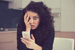 Young brown curly haired shocked anxious person looking at phone message with scared emotion on face. Licensed through Adobe Stock.