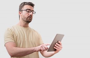 Doubtful man looking at his digital tablet isolated on gray background. Image licensed through Adobe Stock.