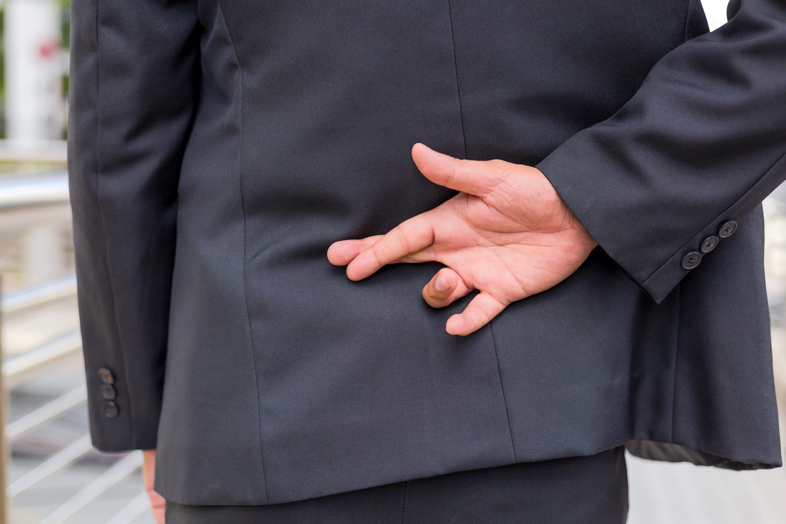 A person in a suit with their hand behind their back and their fingers crossed. Image licensed through Adobe Stock.