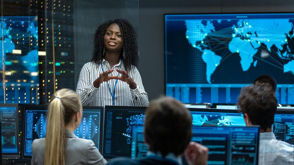 A black woman as Chief Project Engineer Holds Briefing for a Team of Scientists that are Building Machine Learning System. Displays Show Working Model of Neural Network. Image licensed through Adobe Stock.