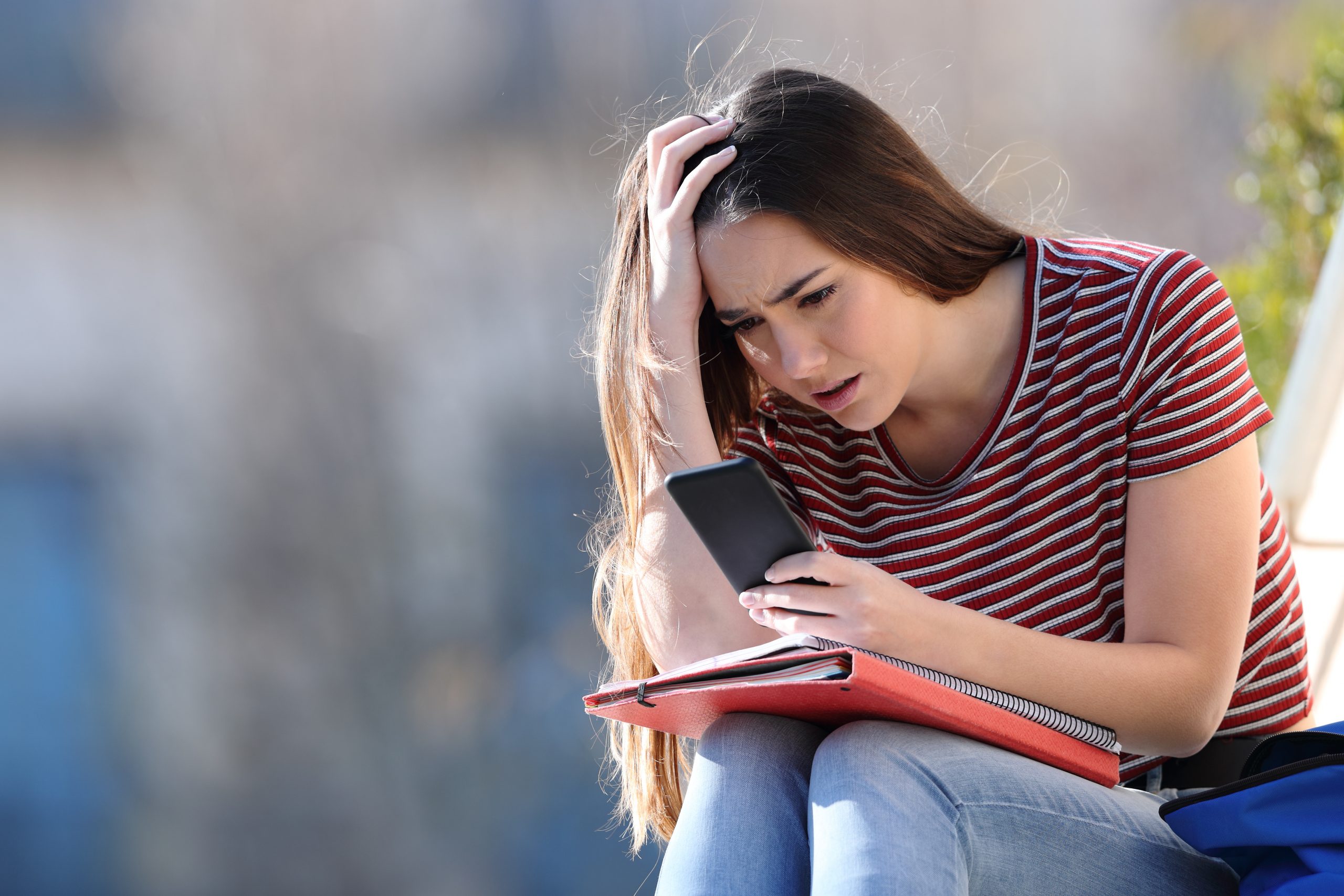 Worried student checking bad news on mobile phone sitting in a campus. Image licensed from Adobe Stock.