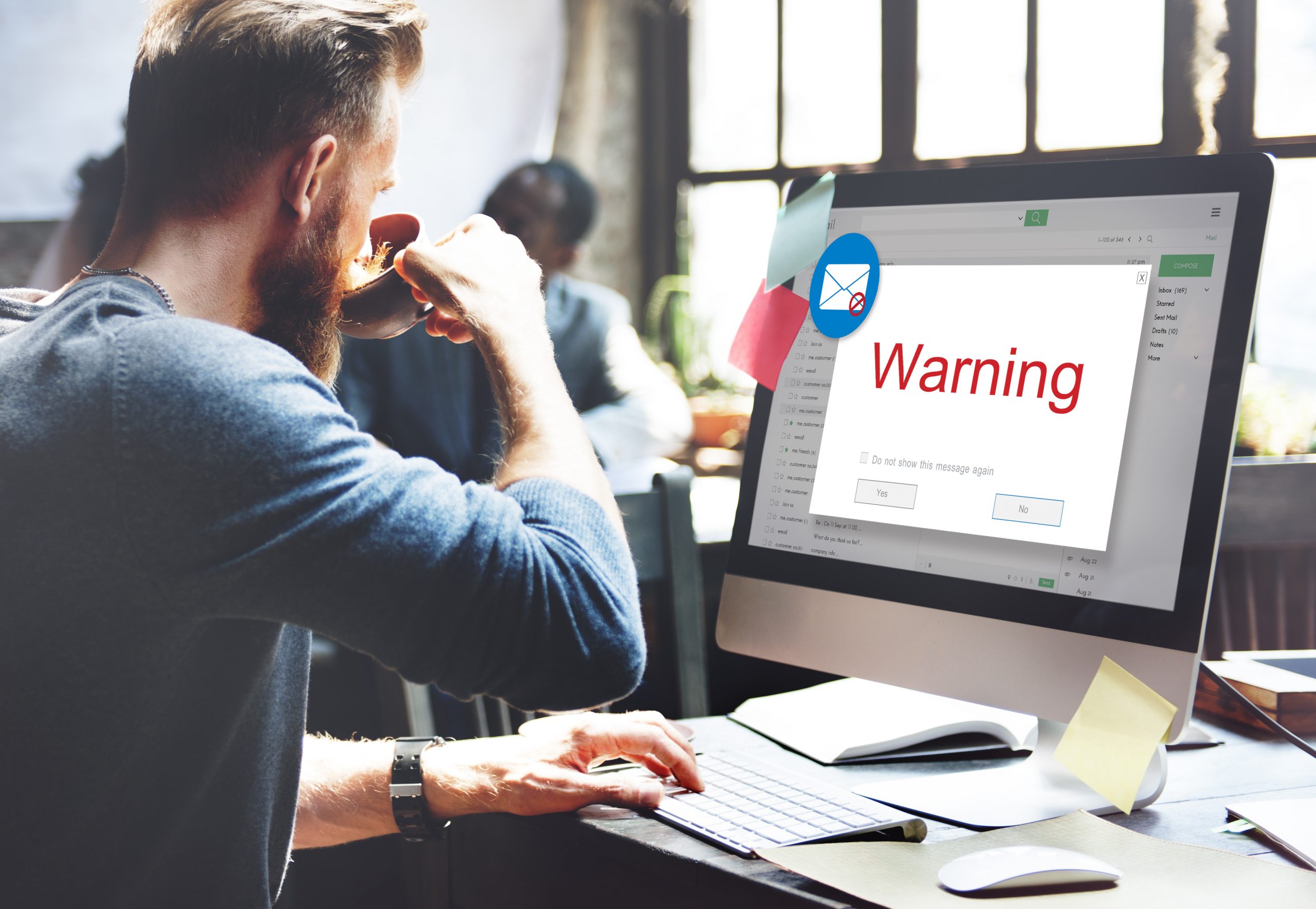 A man drinking coffee at his desk in an office when a warning pop up comes up on his computer screen. Image licensed through Adobe Stock.