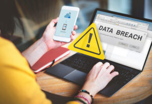 A white person using a laptop on a desk. the laptop has text that reads "Data breach" on the screen with a yellow triangle warning sign displaying an exclamation point imposed over the image. The person is also holding a cell phone with symbols indicating a breach. Image licensed through Adobe Stock.