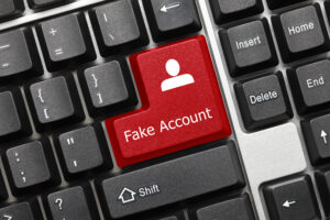 A computer keyboard with black keys and the enter key replaced with a red button displaying white text saying "Fake Account" and a small generic icon representing a person. Image licensed through Adobe Stock.